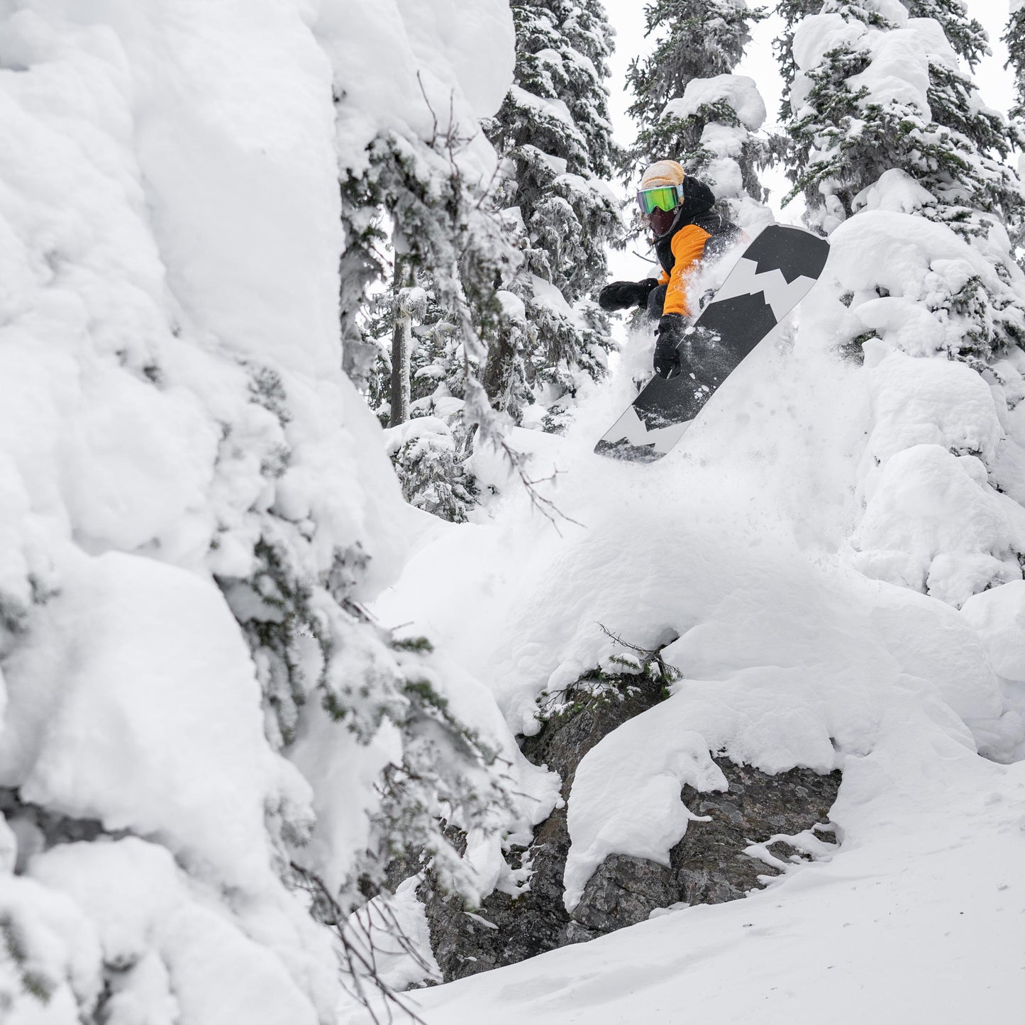 snowboarder riding the weston hatchet snowboard and dropping a cliff grabbing in between his snowboard bindings into deep powder in a snowy forest