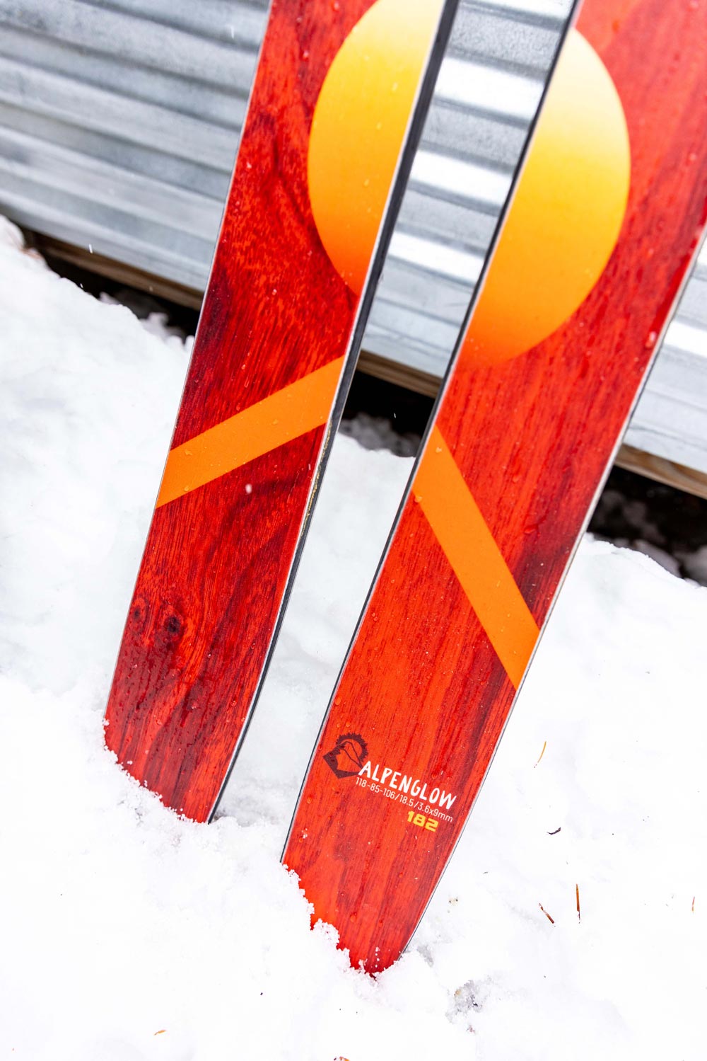 close up of the tails of the weston alpenglow skis stuck in the snow and leaning up against a garage door