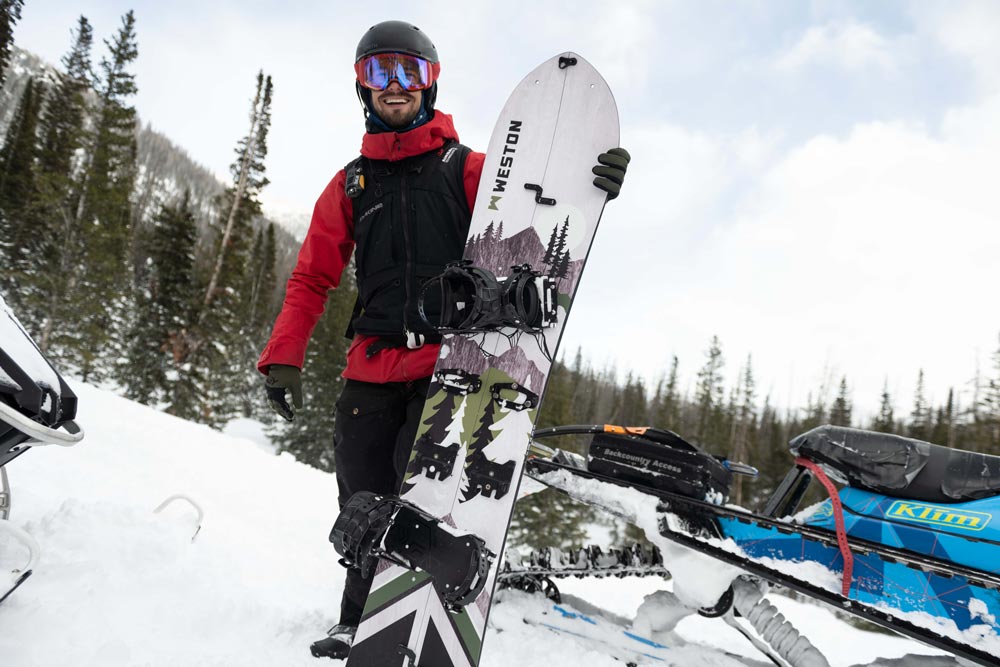 Smiling splitboarder in front of a snowmobile holding a weston backwoods carbon splitboard