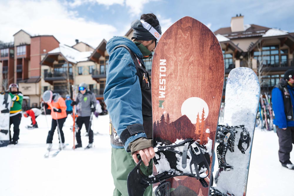 Snowboarder at the base of a ski resort holding the Weston Backwoods snowboard