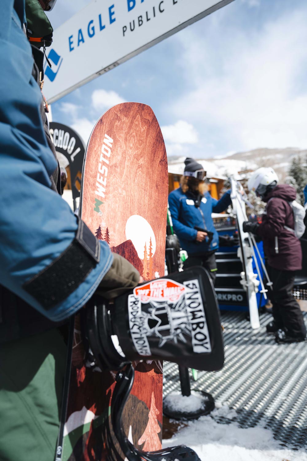 Snowboarder standing in the lift line at a ski resort holding the Weston backwoods snowboard with a ski instructor in the background