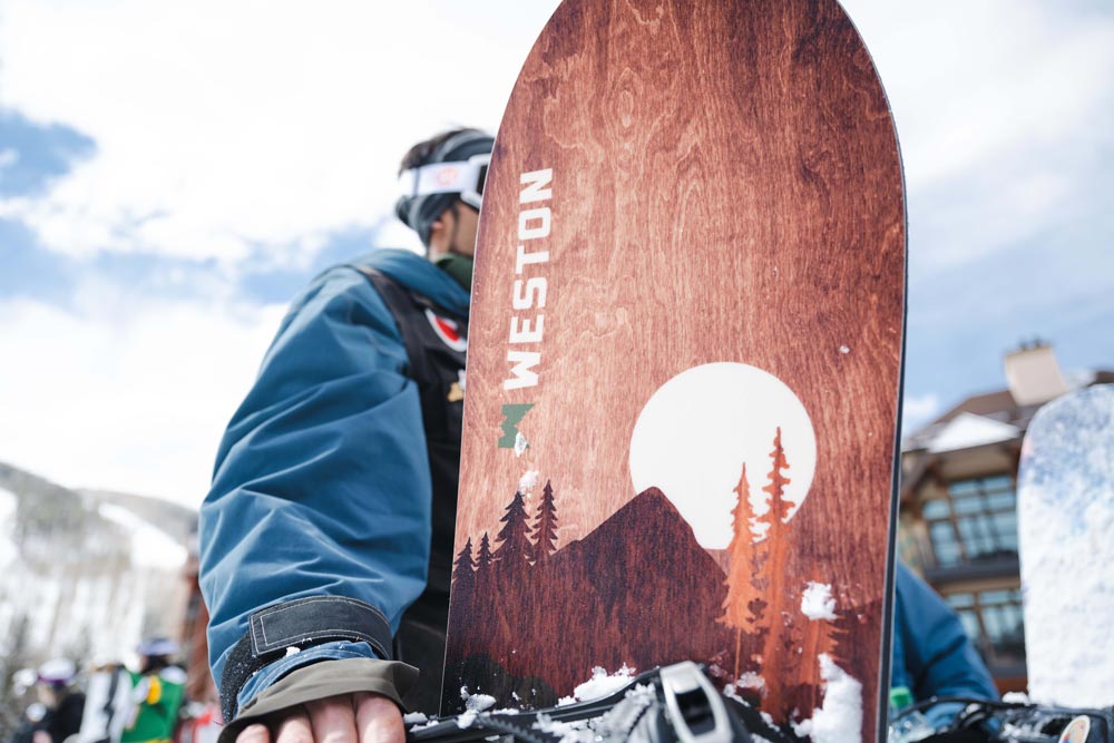 Snowboarder standing in the lift line at a ski resort holding the Weston backwoods snowboard. Close up view of the wood grain graphic featuring a mountaintop and trees