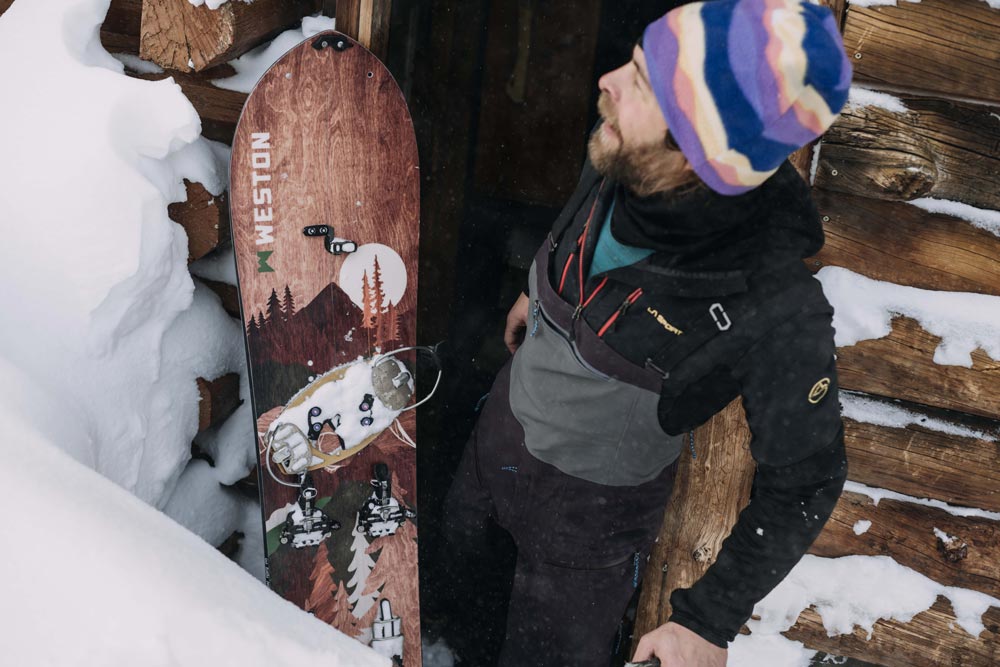 Splitboarder with a purple and yellow kit beanie wearing backcountry bibs leaning outside of a mountain cabin with the weston backwoods splitboard up against the wooden walls