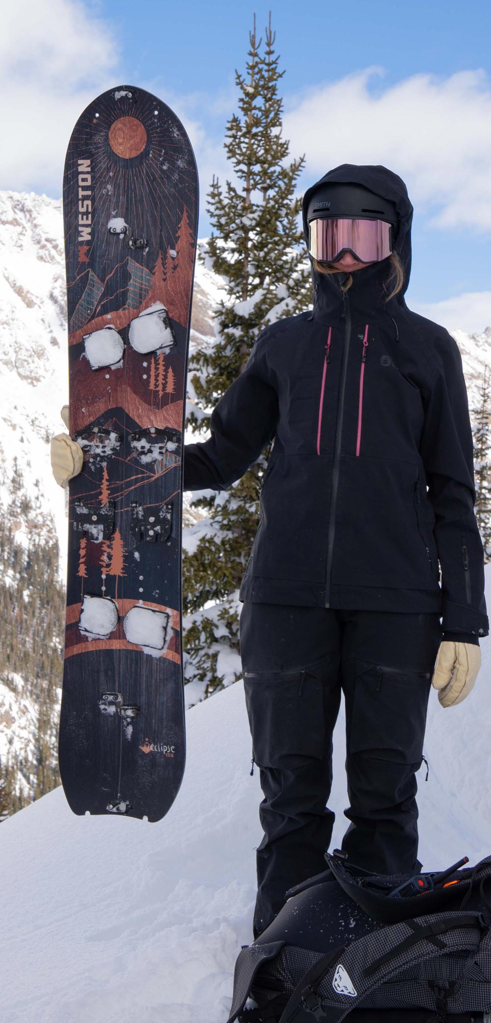 splitboarder wearing ski goggles holding up the weston eclipse snowboard in the backcountry with a tall pine tree in the background