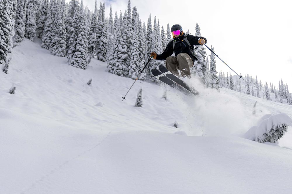 skier on the weston grizzly skis jumping off a snowy mound with snow covered trees in the background