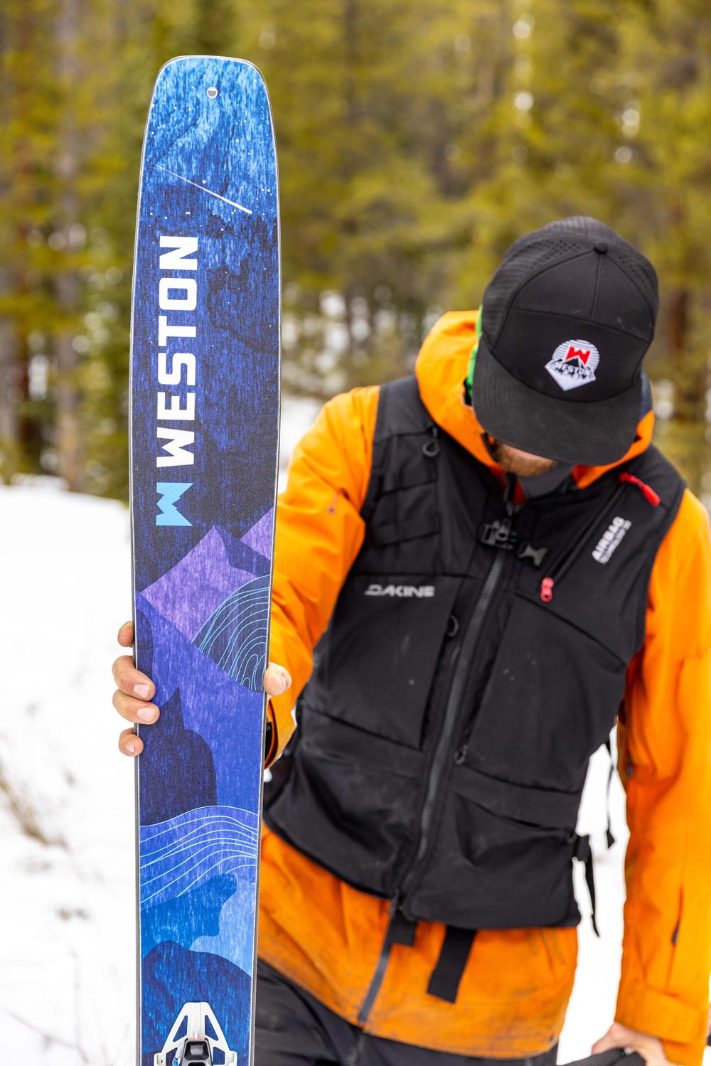 skier wearing a black vest and orange jacket holding a weston skyline ski with a forest background