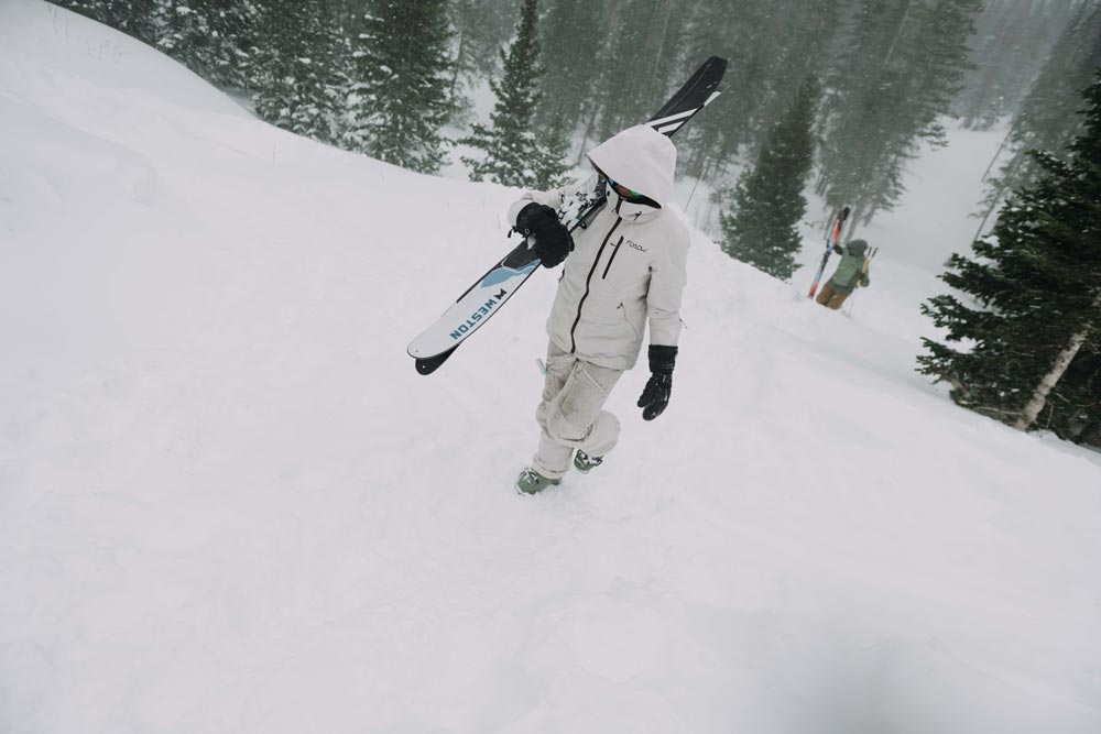 backcountry skier in a white jacket and snowpants walking through a snowy forest holding a pair of weston summit carbon skis perched on her shoulder