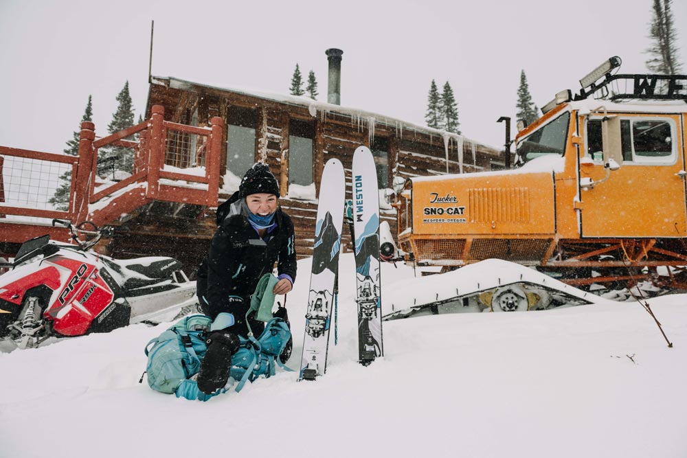 backcountry skier with pair of weston summit carbon skis stuck in the snow in front of the yellow weston tucker snowcat with a wooden backcountry hut in the background