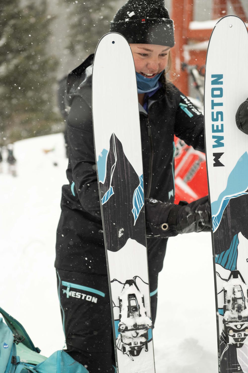 skier in a black beanie and black jacket holding a pair of weston summit carbon skis in the snow in front of a backcountry hut