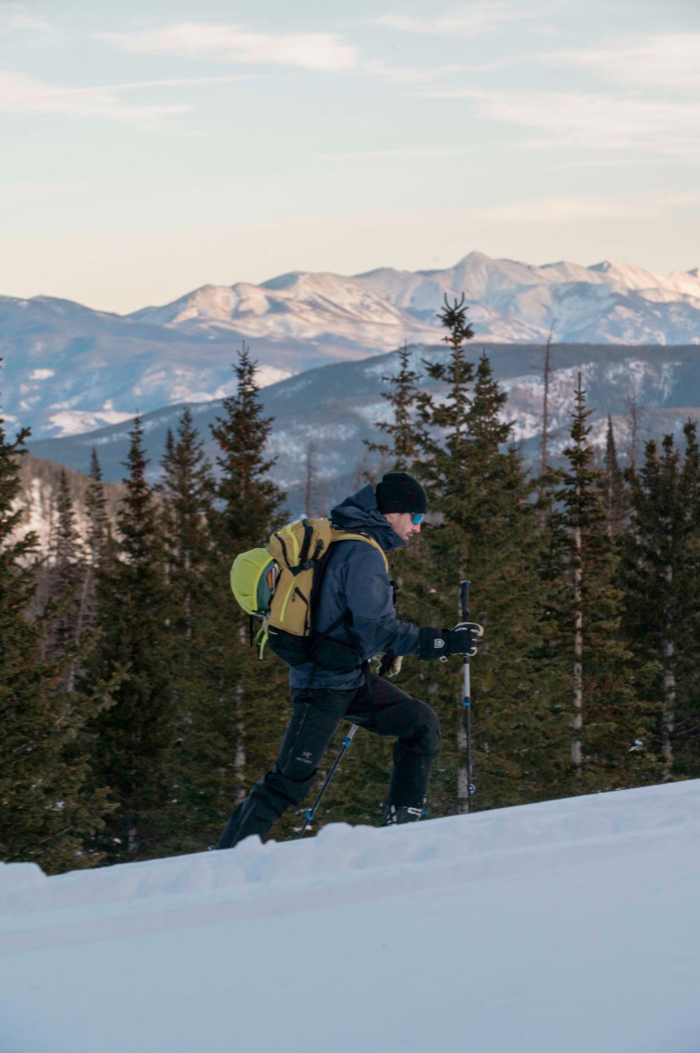 backcountry skier wearing a yellow pack touring up a snowy mountain on the weston summit skis with pine trees in the background