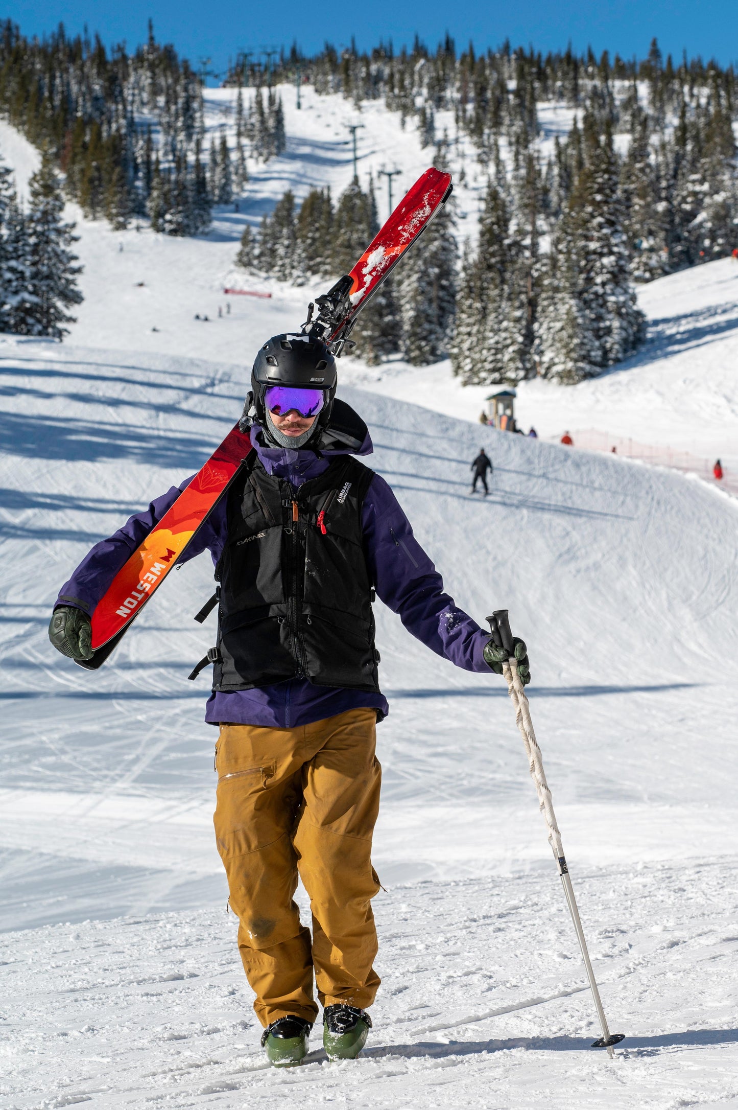skier with a helmet and goggles wearing a black vest and walking with a pair of weston alpenglow skis on his shoulders with a ski resort in the background