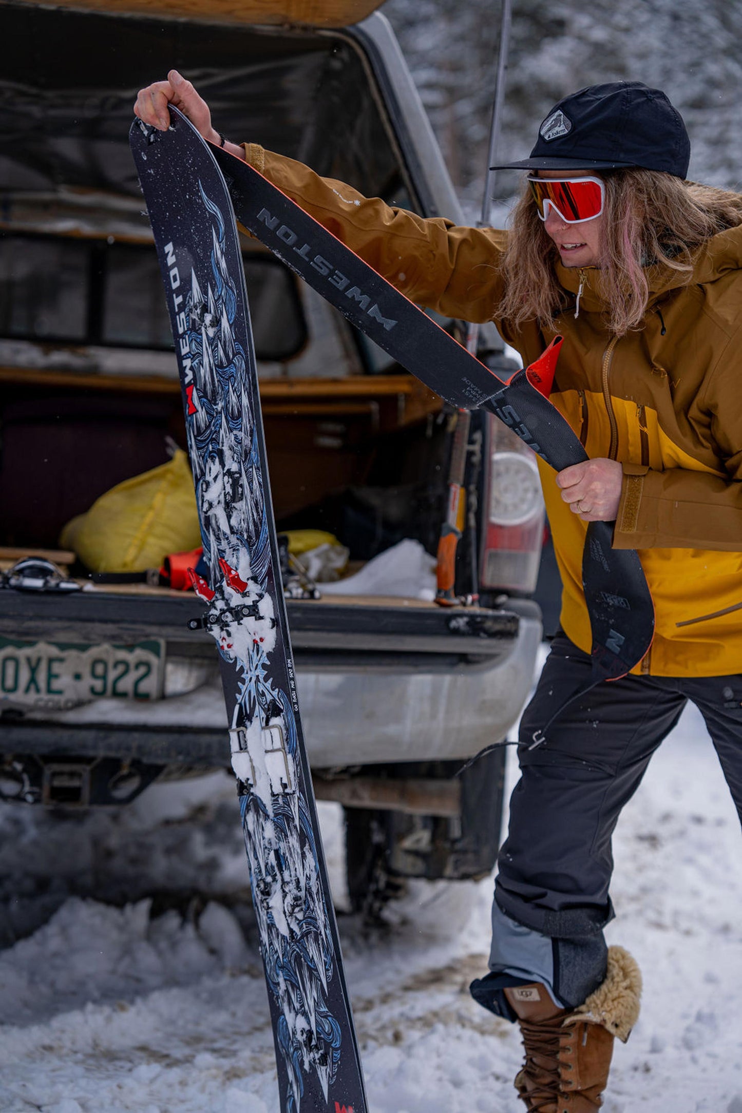 Woman in winter clothing putting backcountry touring skins on one half of the weston dream machine splitboard next to a vehicle in a snowy setting