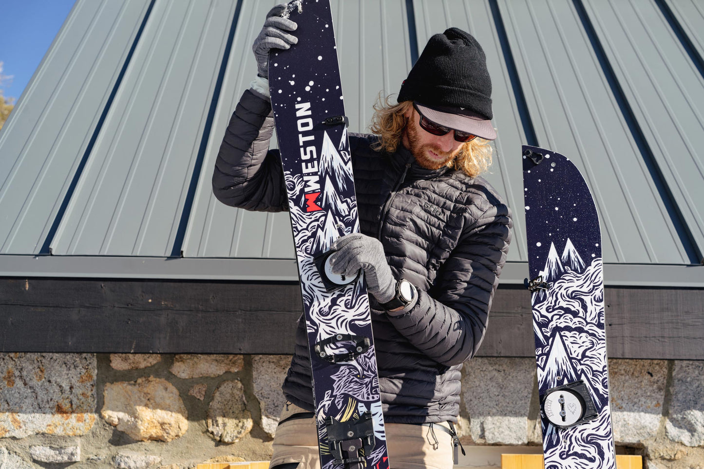 Splitboarder holding two halves of the Weston Gnarnia Splitboard with mountain designs against a building and stone wall background