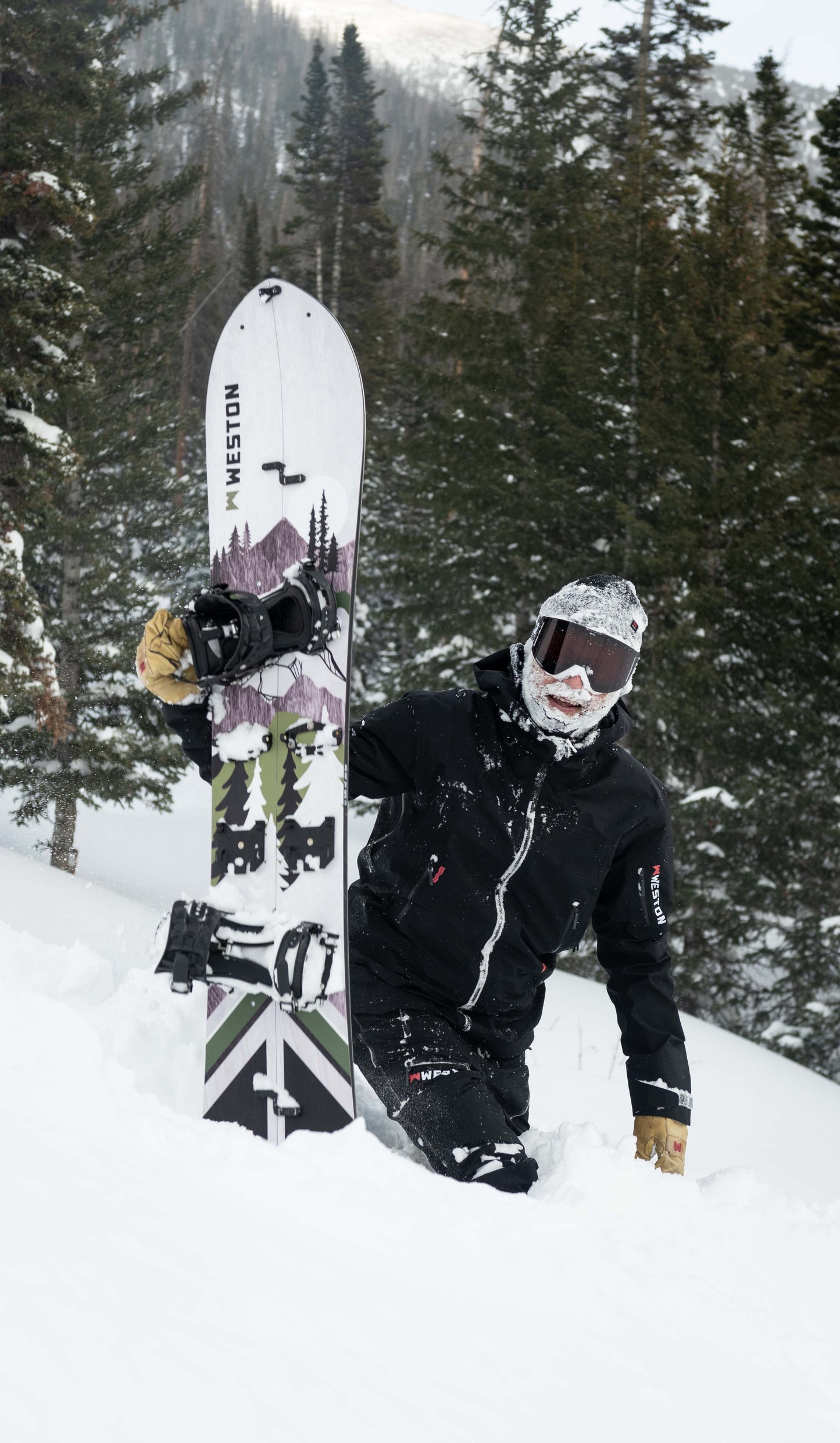 Splitboarder with a face full of snow holding the weston backwoods carbon splitboard in a deep powdery landscape with trees and mountains.