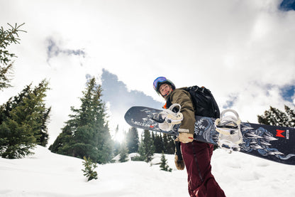 Person holding a weston dream machine snowboard in a snowy landscape with trees and mountains.
