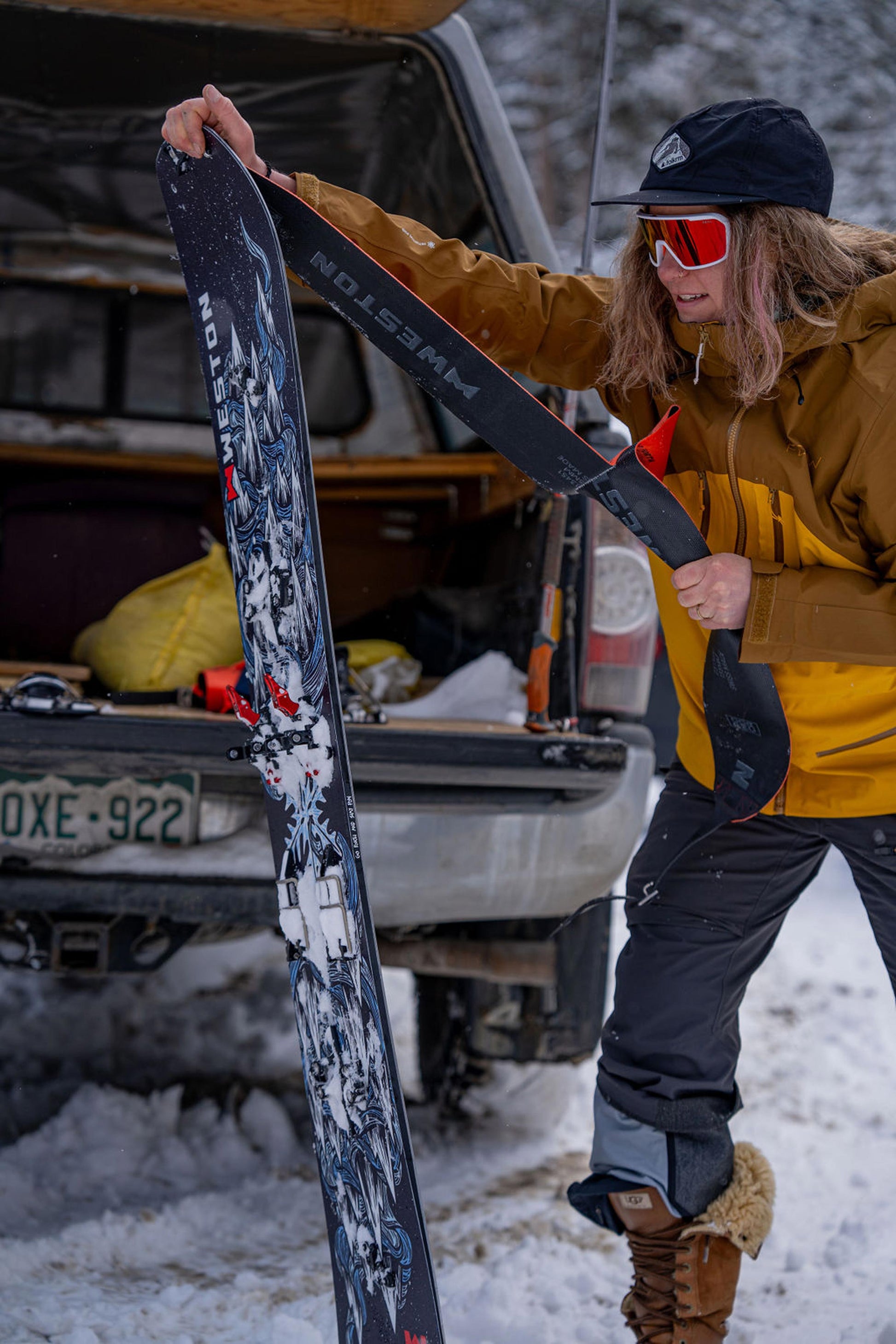 Woman in winter clothing putting backcountry touring skins on one half of the weston dream machine splitboard next to a vehicle in a snowy setting