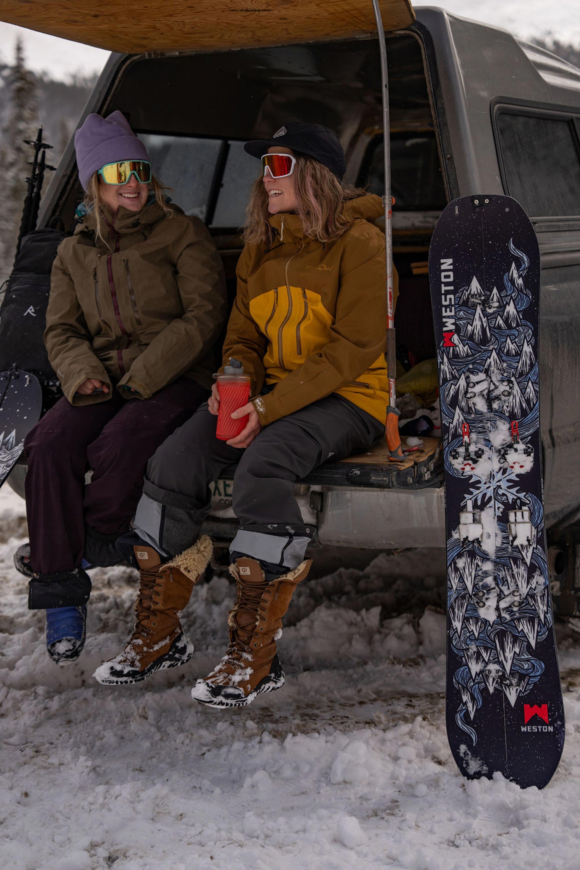 Two splitboarders sitting in the back of a van with the weston dream machine splitboard board leaning against it, in a snowy outdoor setting.